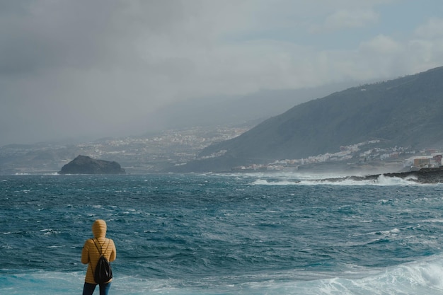 Donna con giacca gialla che guarda l'oceano feroce e aspetta la tempesta