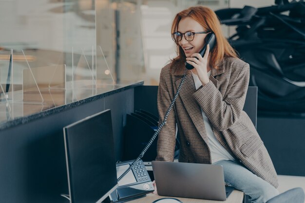 Donna con gli occhiali che lavora in ufficio indossando abiti casual, parlando al telefono mentre è seduta sulla sua scrivania, ambiente d'ufficio moderno e confortevole, sfondo sfocato. Impiegato felice al concetto di lavoro