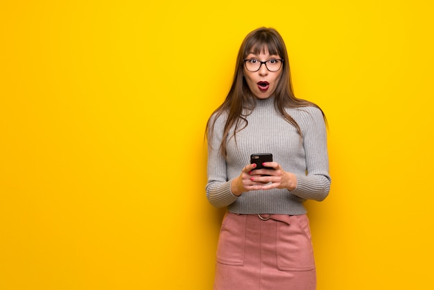 Donna con gli occhiali oltre muro giallo sorpreso e inviando un messaggio