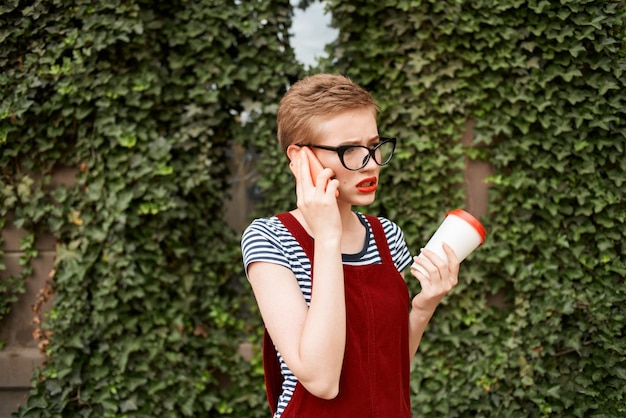 Donna con i capelli corti con gli occhiali per strada parlando al telefono tazza di caffè