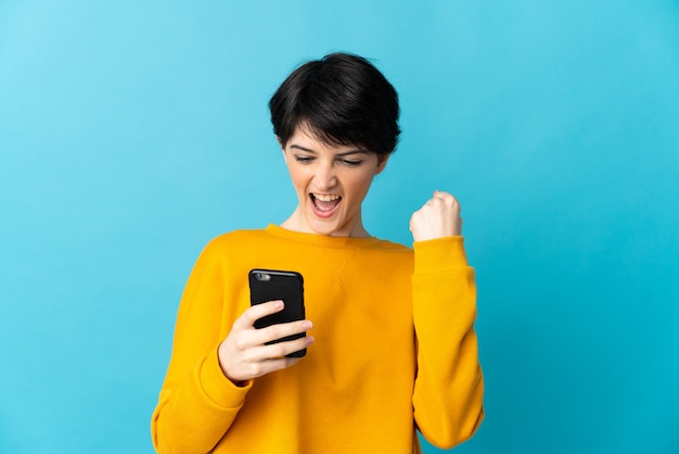 Donna con i capelli corti sopra isolato utilizzando il telefono cellulare e facendo il gesto di vittoria