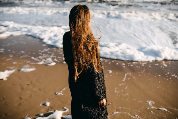 Donna con i capelli lunghi, che indossa un cappotto, in piedi in riva al mare, guardando le onde spumeggianti