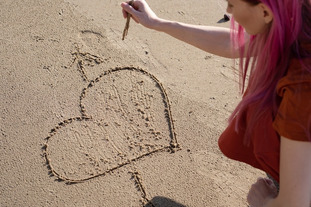 donna con i capelli rosa disegna un cuore con un bastone sulla sabbia