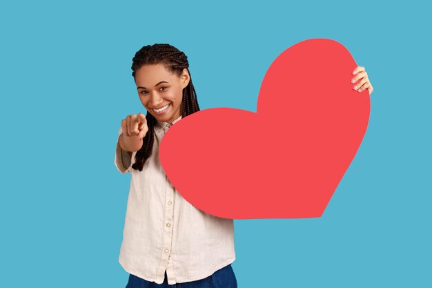 Donna con i dreadlocks che tiene in mano un grande cuore di carta rossa e indica l'amore e il romanticismo della fotocamera