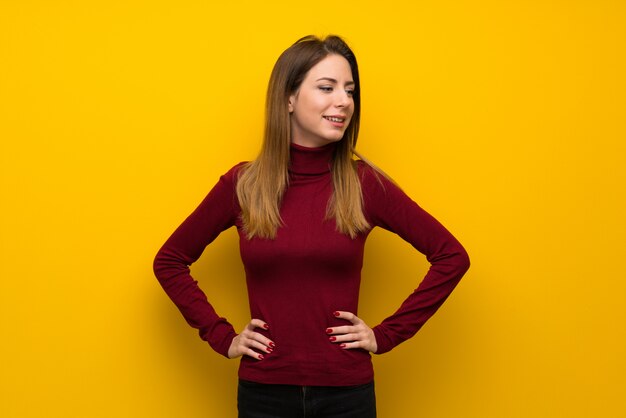 Donna con il collo alto sopra la parete gialla che posa con le armi all&#39;anca e sorridere