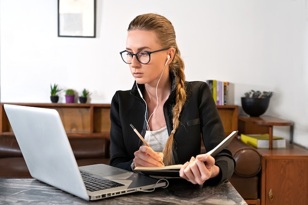 Donna con il computer portatile delle cuffie della bacchetta degli occhiali che studia funzionante in linea