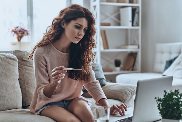 Donna con influenza. Giovani donne malate che tengono in mano un termometro e usano il computer mentre sono sedute sul divano di casa