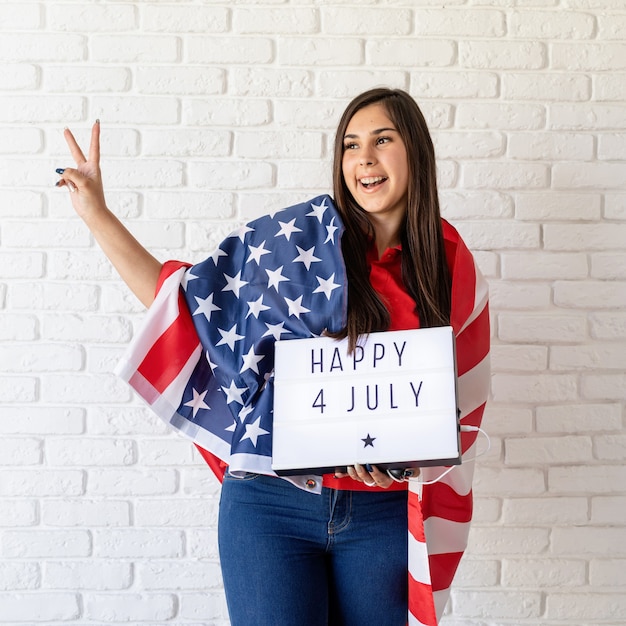 Donna con la bandiera americana che tiene il lightbox con le parole felice 4 luglio e che mostra il segno del pezzo