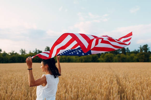 Donna con la bandiera americana nel campo di grano al tramonto il giorno dell'indipendenza di luglio e il concetto di raccolta