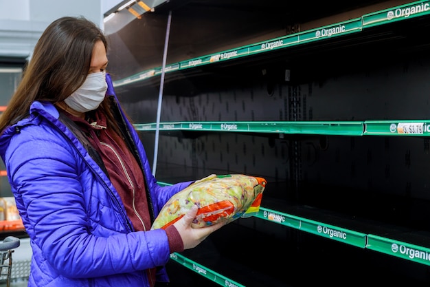 Donna con la maschera che tiene l'ultima borsa di insalata al supermercato con gli scaffali vuoti.