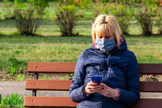 Donna con la maschera protettiva che utilizza Smart Phone nel parco, concetto di coronavirus