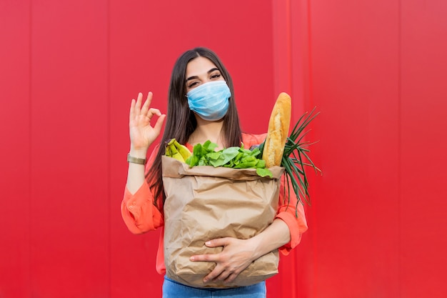 Donna con la mascherina medica che giudica un sacchetto della spesa pieno di alimento fresco. Giovane donna con un sacchetto della spesa durante il covid 19, pandemia di coronavirus.
