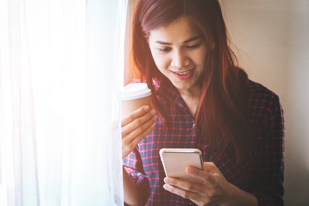 Donna con la tazza di carta caffè utilizzando il telefono cellulare dalle finestre