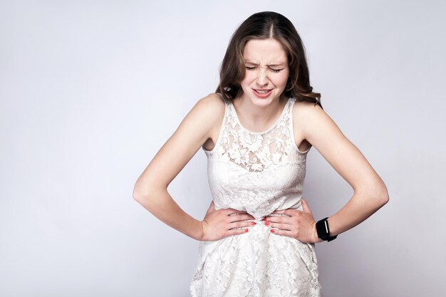 Donna con lentiggini e vestito bianco e orologio intelligente con mal di stomaco su sfondo grigio argento