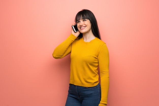 Donna con maglione giallo sul muro rosa mantenendo una conversazione con il telefono cellulare