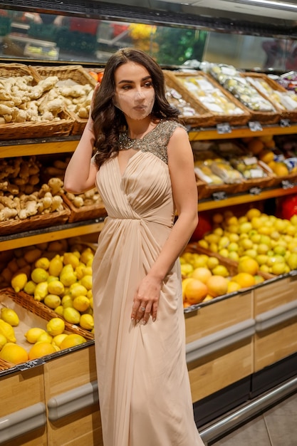 Donna con maschera che fa la spesa in sicurezza durante la pandemia di coronavirus in un negozio di alimentari rifornito. bruna compra frutta, verdura al supermercato. Mancanza di prodotti freschi. Abito da sera scintillante donna