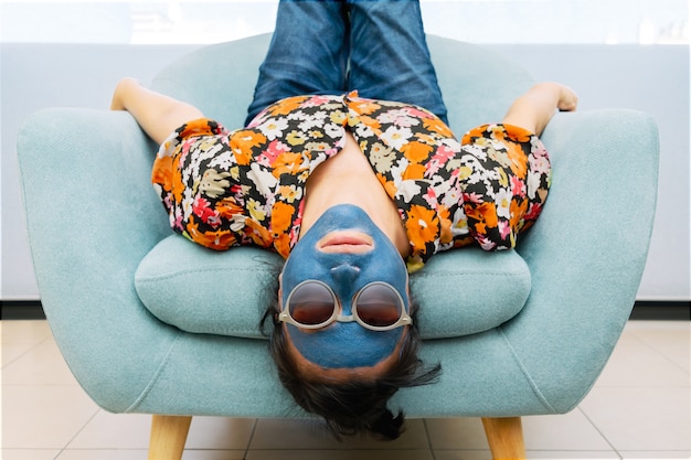 Donna con maschera di bellezza blu e occhiali da sole in una poltrona turchese seduto a testa in giù