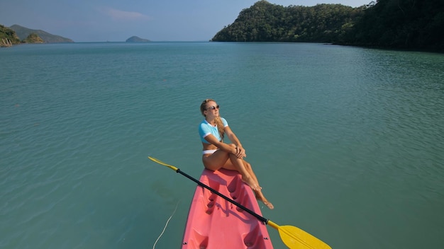 Donna con occhiali da sole righe rosa canoa di plastica lungo l'acqua di mare contro verdi colline e cielo blu