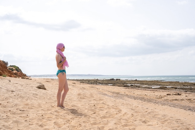 Donna con sciarpa rosa per cancro guardando il mare in bikini dalla spiaggia