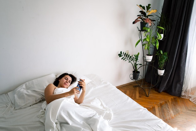 Donna con smartphone a letto con lenzuola bianche