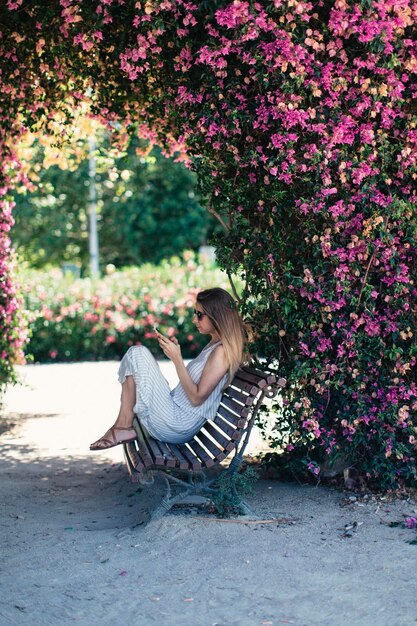 Donna con telefono cellulare seduto sulla panchina nel parco sotto fiori rosa