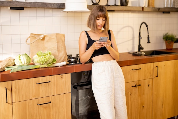 Donna con telefono e generi alimentari in cucina a casa