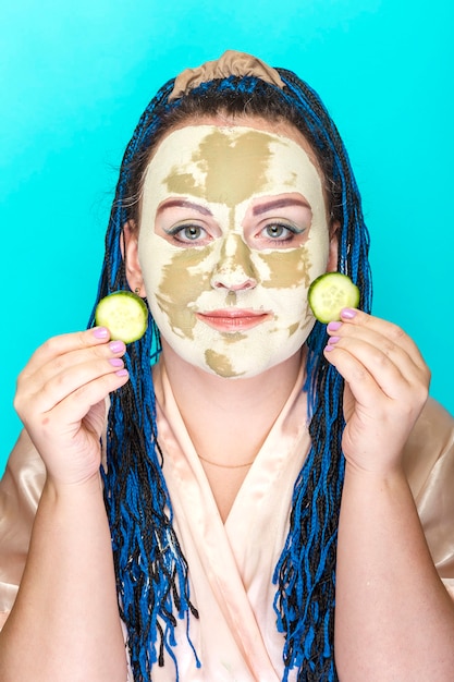 Donna con trecce afro blu faccia in una maschera di argilla verde con cerchi di cetriolo nelle sue mani su sfondo blu Foto verticale