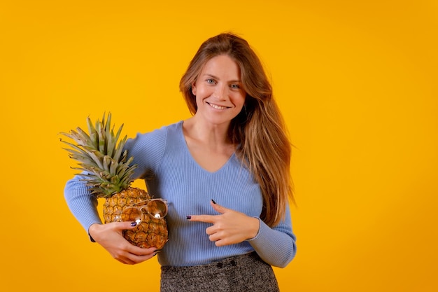 Donna con un ananas in occhiali da sole in uno studio su uno sfondo giallo che punta all'ananas