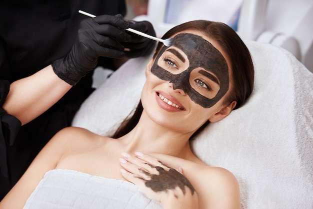 Donna con un bel sorriso che riceve un trattamento viso con maschera al carbonio dall'estetista nella spa, cliente femmina di beauty cllinic avente procedure per il viso