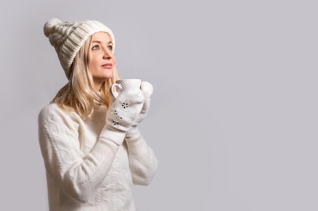 Donna con un caldo maglione bianco di lana con bubo e guanti tiene la tazza sorride e alza lo sguardo sognando