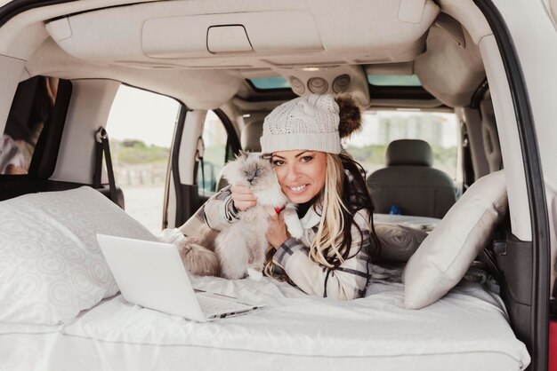 Donna con un cappello bobble che utilizza il computer portatile mentre si trova con il gatto nel rimorchio del camper e guarda la fotocamera Stile di vita dei nomadi digitali