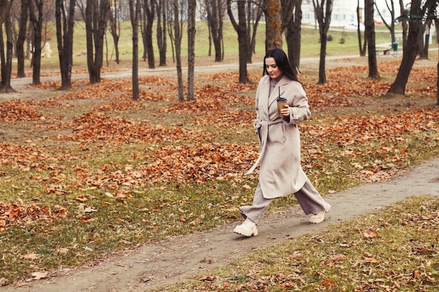 Donna con un cappotto grigio che cammina nel parco autunnale con una tazza di bevanda calda