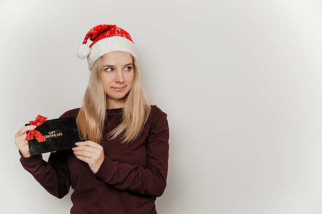 Donna con un maglione bordeaux e un cappello rosso da Babbo Natale con in mano un buono regalo nero su sfondo grigio