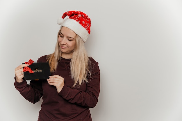 Donna con un maglione bordeaux e un cappello rosso da Babbo Natale con in mano un buono regalo nero su sfondo grigio