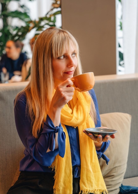 Donna con una camicetta blu e una sciarpa gialla che beve caffè al bar Pensando all'Ucraina