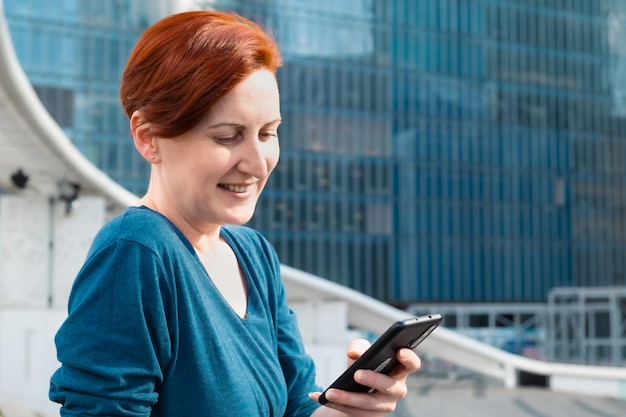 Donna con una camicia blu seduta in un quartiere commerciale della città e guarda lo schermo dello smartphone