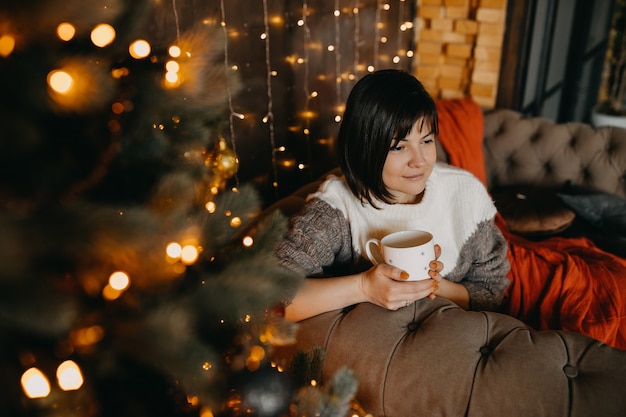 Donna con una tazza di caffè o tè seduto sul divano accanto a un albero di Natale