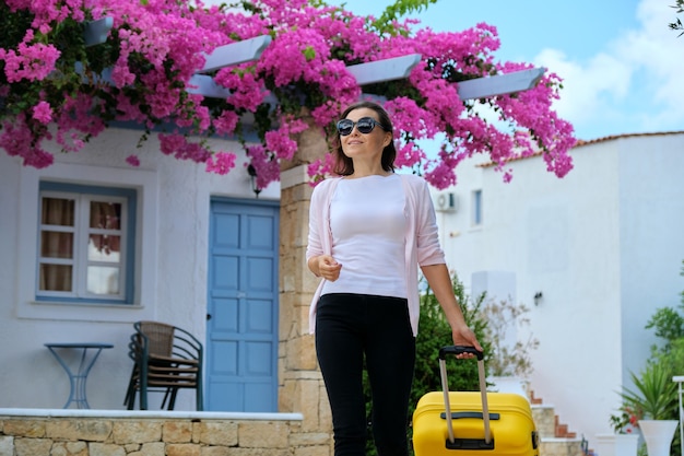 Donna con valigia che cammina all'aperto attraverso il territorio dell'hotel termale pittoresco resort sul mare con un bellissimo paesaggio di fiori rosa. Viaggi, vacanze, tempo libero, fine settimana, persone