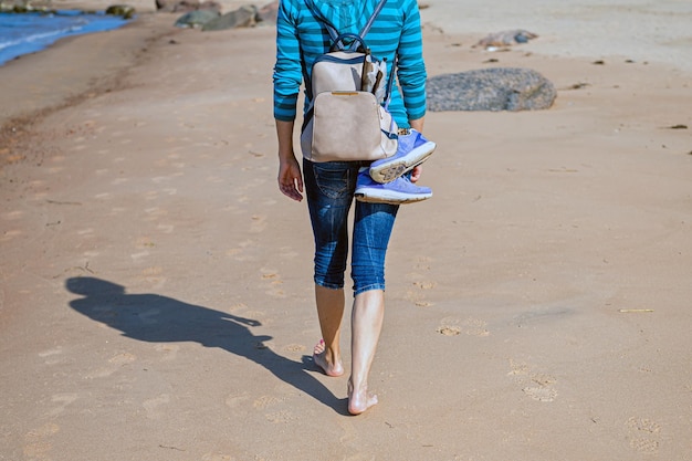 Donna con zaino che cammina a piedi nudi sulla spiaggia durante un primo piano di vista posteriore di una giornata di sole