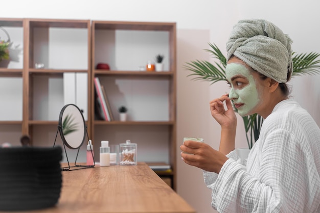 Donna concentrata che applica maschera di argilla fatta in casa sul viso guardando nello specchio del tavolo bella signora