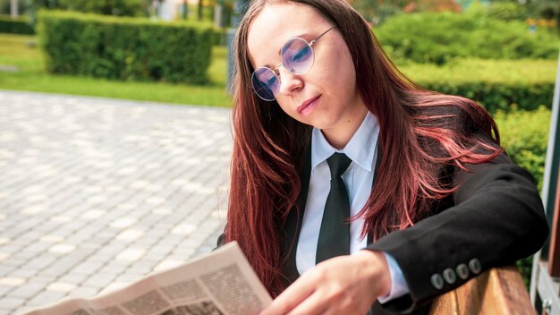 Donna concentrata che legge il giornale nel parco cittadino Giovane donna concentrata in abiti casual e occhiali che legge il giornale mentre è seduta sulla panchina