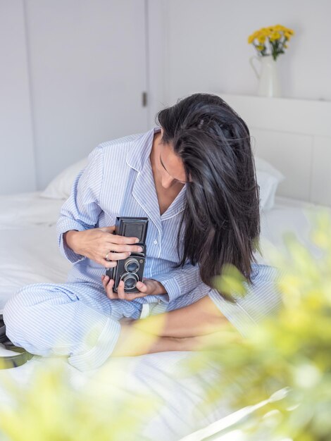 Donna concentrata in pigiama che regola la vecchia macchina fotografica analogica seduta con le gambe incrociate sul letto la mattina