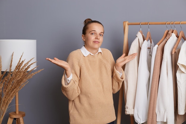 Donna confusa incerta che indossa un maglione lavorato a maglia beige in piedi vicino a scaffali con vestiti che guardano con dubbi pensando e scegliendo abiti alla moda che acquistano nella boutique moderna non possono prendere una decisione