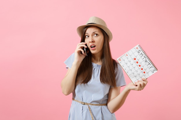 Donna confusa triste che parla al telefono cellulare, tenendo il calendario dei periodi per controllare i giorni delle mestruazioni isolati su sfondo rosa di tendenza brillante. Concetto medico, sanitario, ginecologico. Copia spazio.
