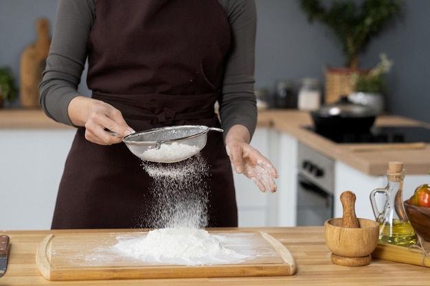 Donna contemporanea in grembiule che setaccia la farina dal tavolo della cucina