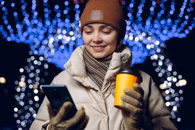 Donna contenta con caffè per andare a usare il cellulare nella città invernale con ghirlande luminose