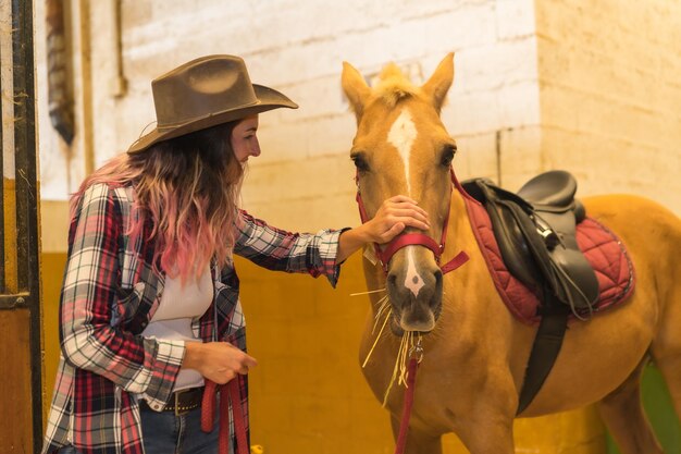 Donna cowgirl dai capelli rosa caucasica che passeggia con un cavallo in una stalla, cappelli da cowboy americani, camicia a quadri rosa e jeans