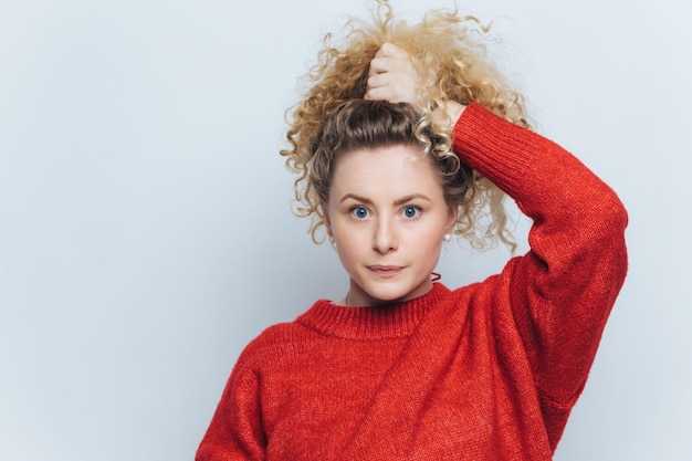 Donna curiosa con i capelli ricci voluminosi in un maglione rosso vibrante mano nei capelli guardando pensierosamente da parte