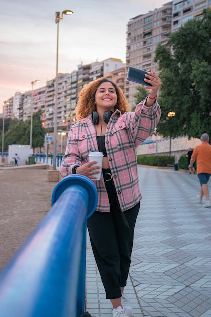 Donna curvy con capelli ricci prende un selfie con il suo telefono sulla costa al tramonto