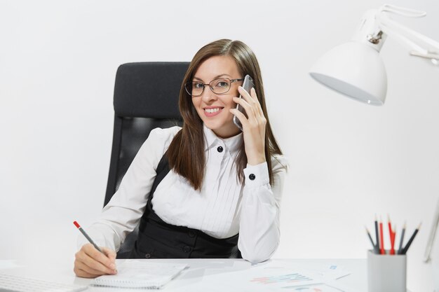 Donna d'affari abbastanza sorridente in tuta seduta alla scrivania, lavorando al computer contemporaneo con documento in ufficio leggero, parlando al telefono cellulare, conducendo una piacevole conversazione